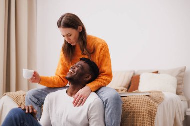Kadın elinde kahve tutuyor ve evde yatak odasında Afrikalı Amerikalı erkek arkadaşıyla eğleniyor.