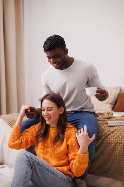 Gülümseyen Afrikalı Amerikalı bir adam kız arkadaşının elini tutuyor ve yatak odasında kahve içiyor.