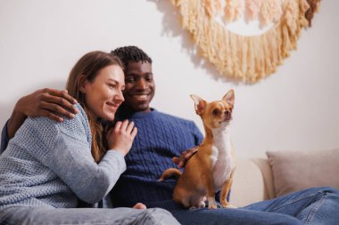 Neşeli, çoklu etnik çeşitlilikte genç bir çift akşamları evdeki kanepede köpek besliyor.