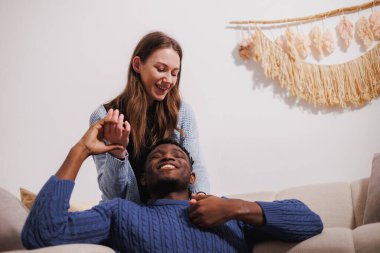 Evdeki koltukta el ele tutuşan gülümseyen çok ırklı çift.