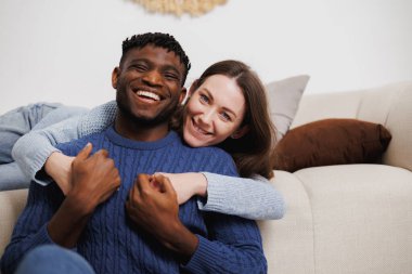 Kameraya bakıp oturma odasında el ele tutuşan gülümseyen, etnik çeşitlilikte genç bir çift.