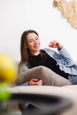 Evdeki kanepede dinlenirken gülümseyen kadın kameraya bakıyor.