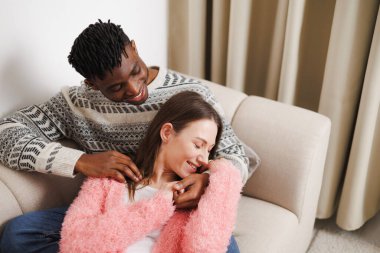 Çok ırklı neşeli bir çift evdeki kanepede sarılıp dinleniyor.