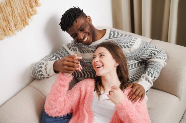 Gülümseyen, etnik çeşitlilikte çiftler evdeki koltukta el ele tutuşuyorlar.