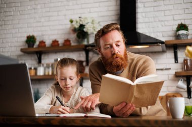 Sakallı baba evde mutfakta online eğitimi olan kızını desteklerken kitap okuyor.