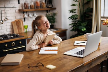 Çevrimiçi ders sırasında dizüstü bilgisayarlar ve mutfaktaki gözlüklerin yanında kitap tutan neşeli ergen çocuk.