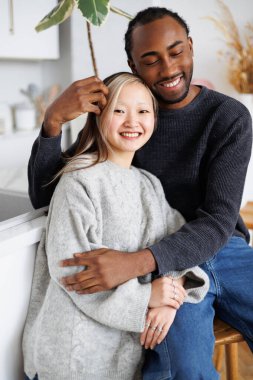 Neşeli Afrikalı Amerikalı adam mutfakta Asyalı kız arkadaşına sarılıyor.