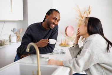 Asyalı kız arkadaşı modern mutfakta konuşurken, elinde kahve tutan gülen Afro-Amerikan bir adam.