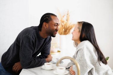 Neşeli, genç, etnik çeşitlilikte bir çift mutfakta kahve içerek vakit geçiriyorlar.