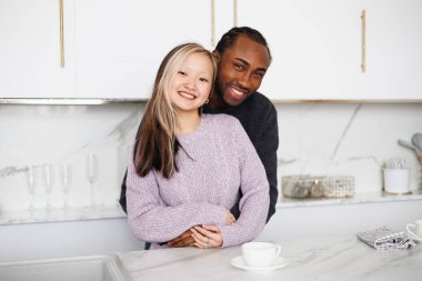 Gülümseyen, etnik çeşitlilikte genç bir çift mutfaktaki kahvenin yanında sarılıp kameraya bakıyor.