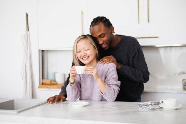 Genç Afro-Amerikalı adam mutfakta gülümseyen Asyalı kız arkadaşına sarılıyor.