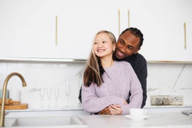 Neşeli, genç, etnik çeşitlilikte bir çift modern mutfakta kahvenin yanında sarılıyor.