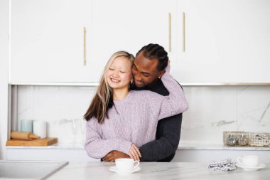 Gülümseyen Asyalı bir kadın. Afro-Amerikan kız arkadaşına sarılıyor. Mutfakta kahvenin yanında.