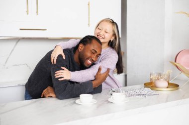 Süveter giyen neşeli genç Asyalı kadın mutfakta kahvenin yanında Afrikalı Amerikalı sevgiliye sarılıyor.