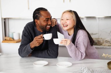 Evde mutfakta konuşurken kahve fincanı tutan pozitif ırklar arası genç bir çift.