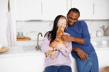 Mutfakta Chihuahua köpeğiyle oynayan gülümseyen çok ırklı genç bir çift.