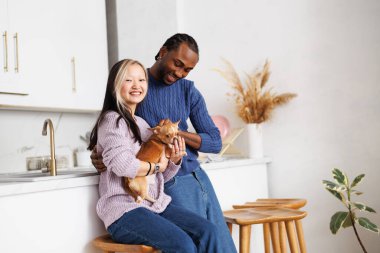 Gülümseyen, etnik çeşitlilikte süveter giyen genç bir çift evde mutfakta chihuahua köpeği tutuyor.