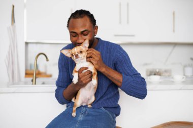 Kazaklı ve kot pantolonlu genç Afro-Amerikan adam mutfakta chihuahua köpeği besliyor.