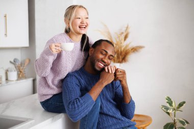 Gülümseyen Afro-Amerikan kazaklı adam mutfakta kahveli Asyalı kız arkadaşının elini tutuyor.