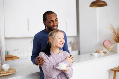 Gülümseyen Afro-Amerikan adam Asyalı kız arkadaşına sarılıyor evde kahve tutuyor.