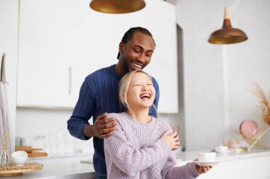 Pozitif Afro-Amerikalı adam Asyalı kız arkadaşına sarılıyor mutfakta kahve tutuyor.
