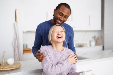 Gülümseyen Afrikalı Amerikalı adam Asyalı kız arkadaşına mutfakta kahveyle sarılıyor.