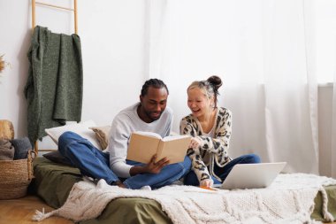 Evde laptopun yanında kitap okuyan çok ırklı bir çift var.