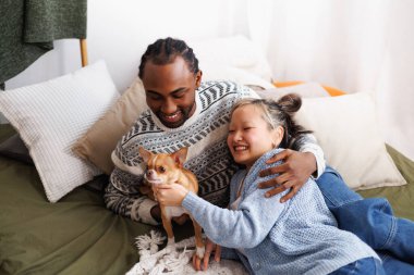 Gülümseyen genç, çok kültürlü bir çift evde yatakta köpekle dinleniyor.