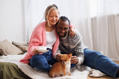 Gülümseyen Asyalı kadın evde Afro-Amerikan erkek arkadaşıyla, evcil hayvanıyla ve kahvesiyle rahatlıyor.