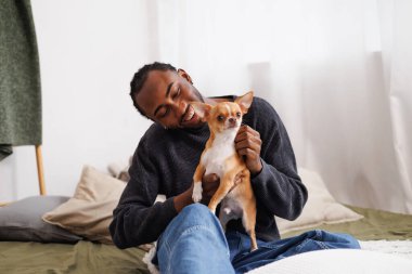 Kapüşonlu tişörtlü Afro-Amerikan bir adam yatak odasında chihuahua köpeği tutuyor.