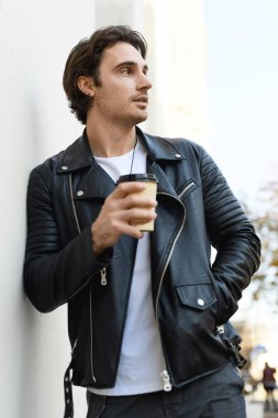 Deri ceketli yakışıklı esmer adam beyaz duvara yaklaşmak için elinde kahve tutuyor.