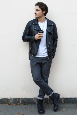 Deri ceketli şık bir adam elinde kahveyle beyaz duvarın yanındaki pantolonun cebini dolduruyor.