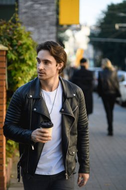Deri ceketli, sırıtan esmer adam sokakta yürürken elinde kahve tutuyor.
