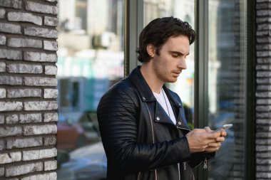 Deri ceketli genç adam binanın yanında cep telefonu kullanıyor.
