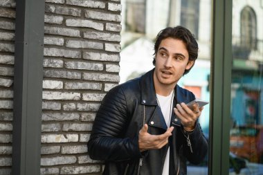 Şehir caddesinde sesli mesaj kaydederken akıllı telefon kullanan deri ceketli yakışıklı bir adam.