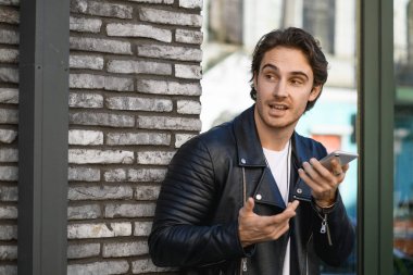 Deri ceketli şık bir adam akıllı telefondan sesli mesaj kaydediyor ve caddeye bakıyor.