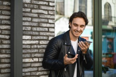 Deri ceketli genç adam binanın yakınındaki akıllı telefondan sesli mesaj kaydediyor.