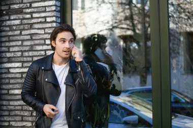 Deri ceketli şık adam şehir caddesindeki binanın yakınında akıllı telefondan konuşuyor.