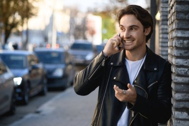 Deri ceketli esmer adam şehir caddesinde yol kenarında cep telefonuyla konuşuyor.