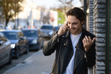 Deri ceketli şık bir adam. Şehir caddesinde bina ve yol kenarında akıllı telefonla konuşurken gülümsüyor.