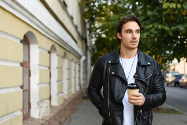 Deri ceketli yakışıklı esmer adam, şehirde yürürken elinde kağıt bardak tutuyor.