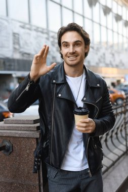 Deri ceketli gülümseyen adam kağıt bardak tutuyor ve şehirde el sallıyor.