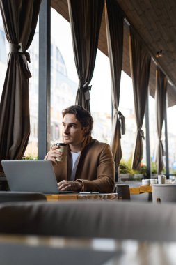 İyi görünümlü bir işadamı kafenin terasında dizüstü bilgisayar ve cep telefonunun yanına gitmek için kahve tutuyor.