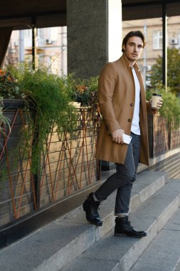 Şık bir işadamı akıllı telefon tutuyor ve binanın yanındaki merdivenlerde kahve içiyor.