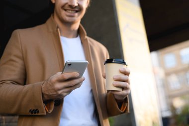 Paltoyla gülümseyen iş adamının elinde içecek ve şehir caddesinde akıllı telefon kullanması görüntüsü.