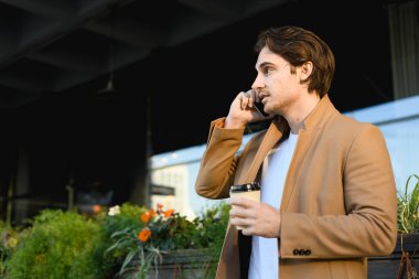 Palto giymiş, elinde kahve olan yakışıklı işadamının şehir caddesinde cep telefonuyla konuşmasının yan görüntüsü.
