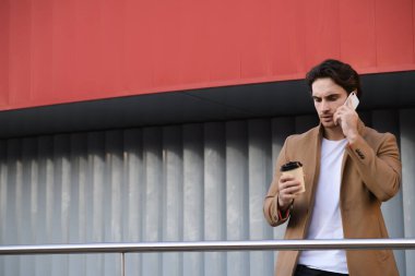 Ceketli ciddi bir işadamı cep telefonuyla konuşuyor ve tek kullanımlık bardağı dışarıda tutuyor.