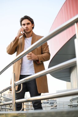 Palto giymiş bir işadamı elinde kahve ile şehir caddesindeki binanın parmaklıklarının yanındaki telefondan konuşacak.