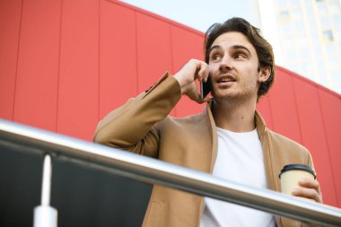 Palto giymiş genç işadamlarının akıllı telefondan konuşmaları ve binanın yakınında içecek bulundurmaları düşük açılı bir bakış açısı.