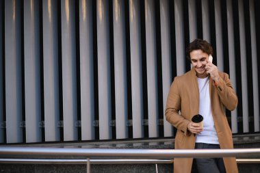 Kahverengi ceketli, gülümseyen müdür elinde kahve ile şehir caddesinde cep telefonuyla konuşuyor.